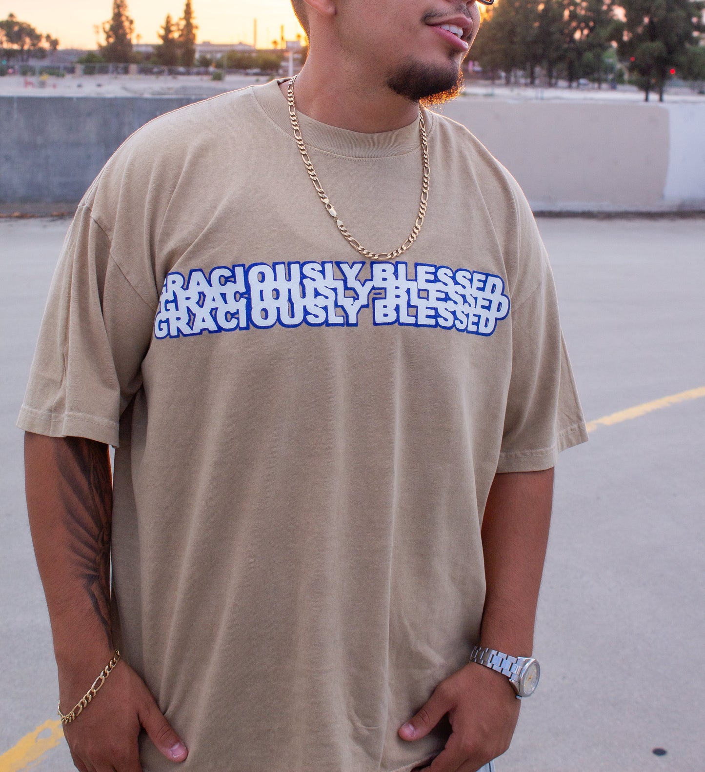 Man wearing a beige t-shirt with 'RIGHTEOUSLY BLESSED' text, sunglasses, and gold chain in an outdoor setting.
