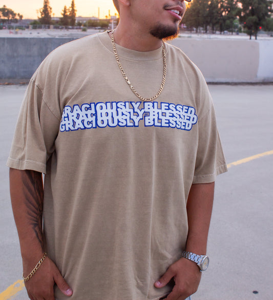 Man wearing a beige t-shirt with 'RIGHTEOUSLY BLESSED' text, sunglasses, and gold chain in an outdoor setting.